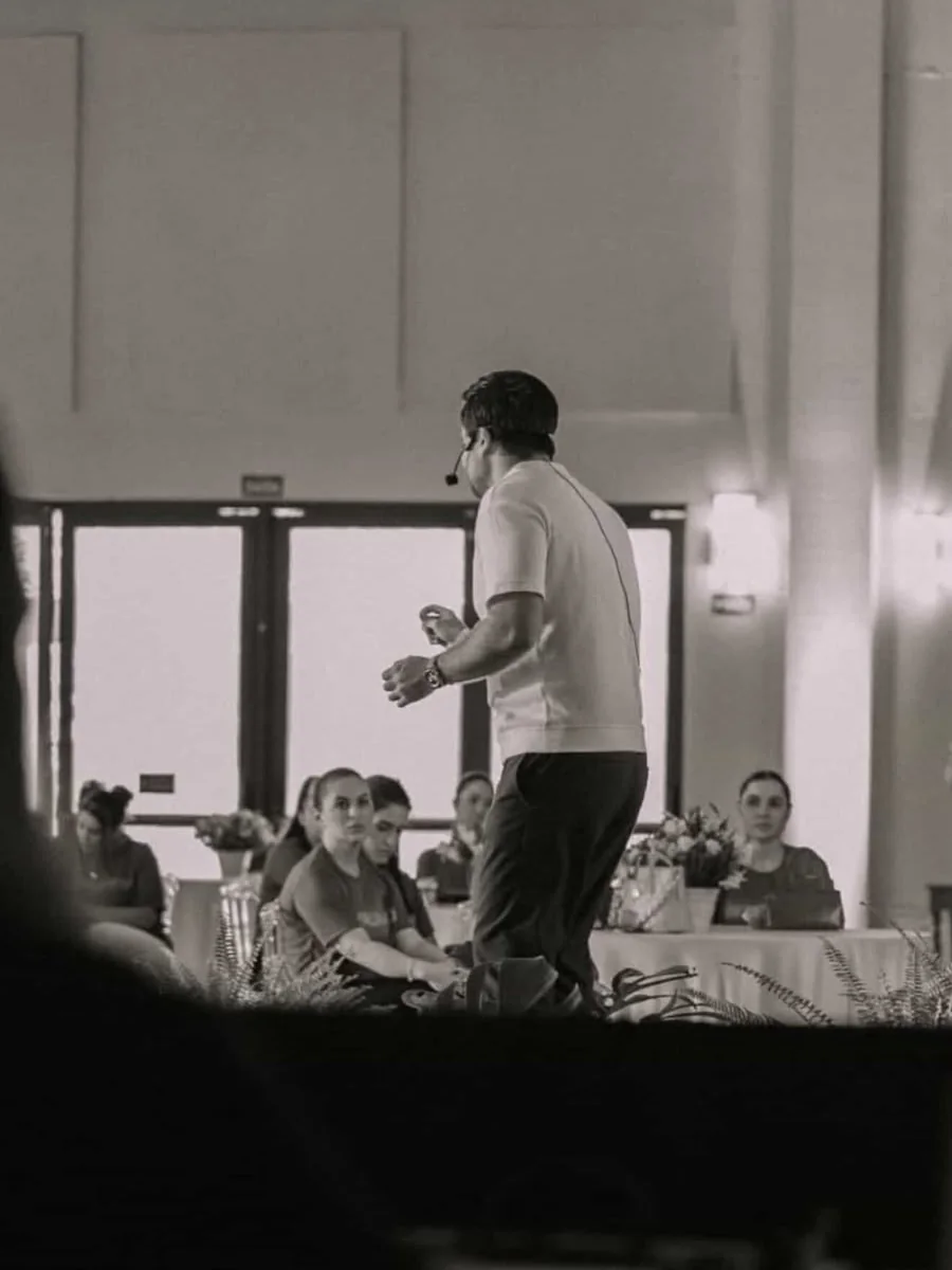 Minoru Raphael de costas, com microfone de cabeça, falando para a plateia, em foto em preto e branco Minoru Raphael de costas, com microfone de cabeça, falando para a plateia, em foto em preto e branco