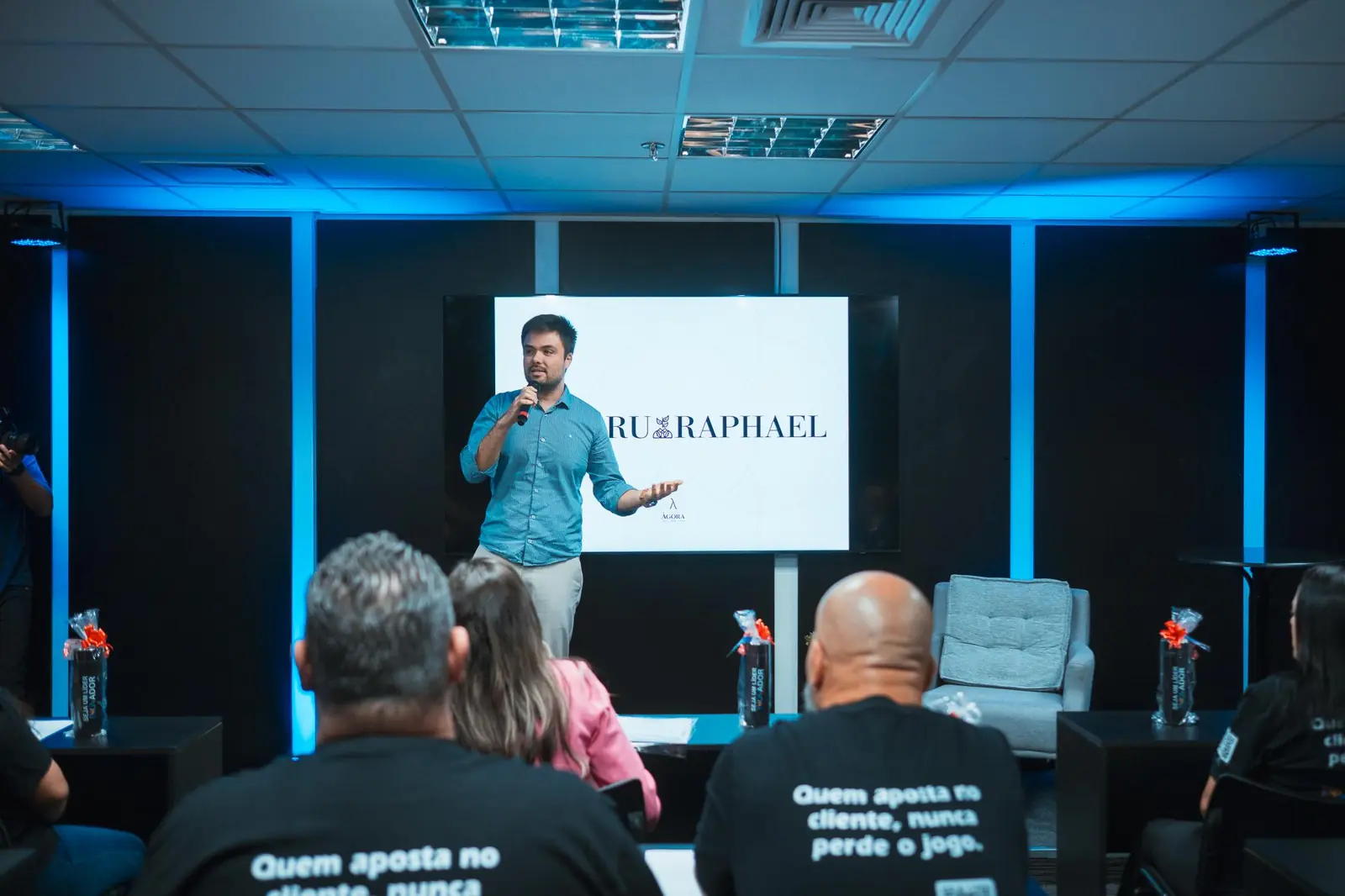 Minoru Raphael de camisa azul e microfone na mão, palestrando diante de um telão, com a plateia de costas em primeiro plano Minoru Raphael de camisa azul e microfone na mão, palestrando diante de um telão, com a plateia de costas em primeiro plano