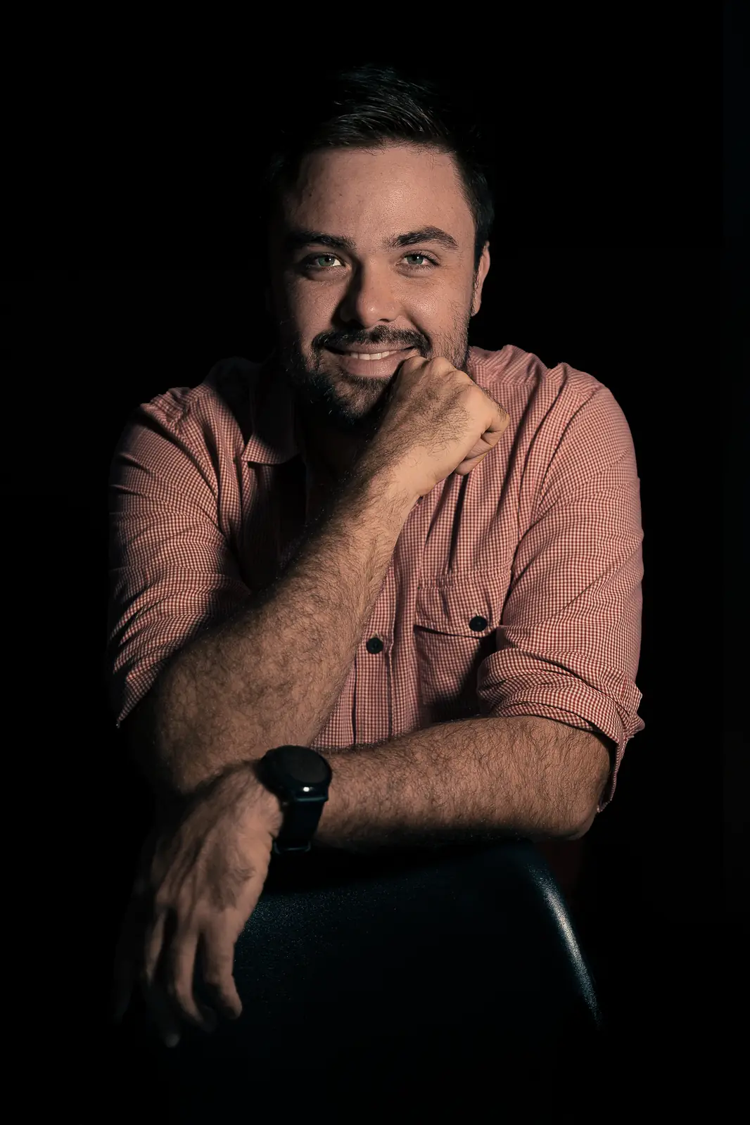 Minoru Raphael sorrindo, com o queixo apoiado na mão, de camisa xadrez, em retrato de fundo preto Minoru Raphael sorrindo, com o queixo apoiado na mão, de camisa xadrez, em retrato de fundo preto