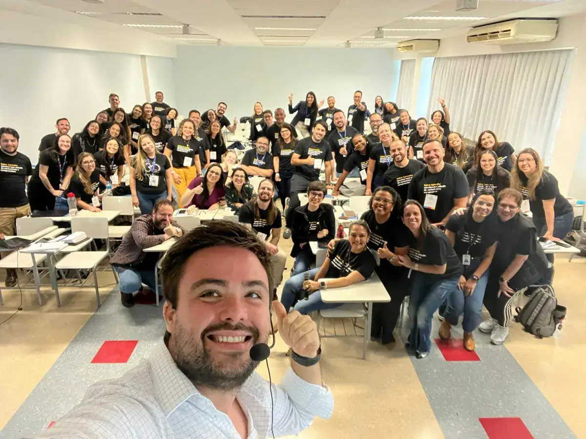 Minoru Raphael de camisa branca e microfone de cabeça, num selfie com uma turma de treinamento sorrindo, de camiseta preta, numa sala de aula Minoru Raphael de camisa branca e microfone de cabeça, num selfie com uma turma de treinamento sorrindo, de camiseta preta, numa sala de aula