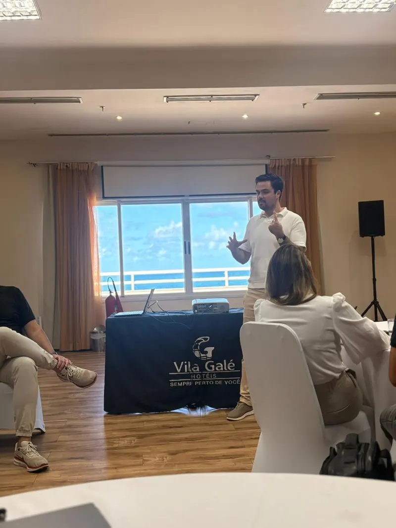 Minoru Raphael de camisa polo branca, gesticulando, durante uma palestra numa sala com janela para o mar Minoru Raphael de camisa polo branca, gesticulando, durante uma palestra numa sala com janela para o mar