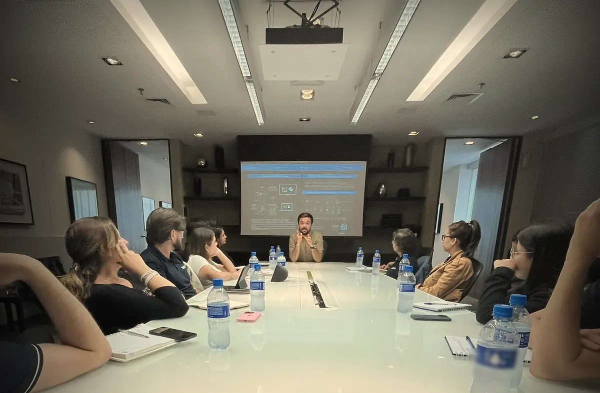 Minoru Raphael falando na ponta de uma mesa de reunião, diante de um telão, com a equipe sentada ao redor Minoru Raphael falando na ponta de uma mesa de reunião, diante de um telão, com a equipe sentada ao redor